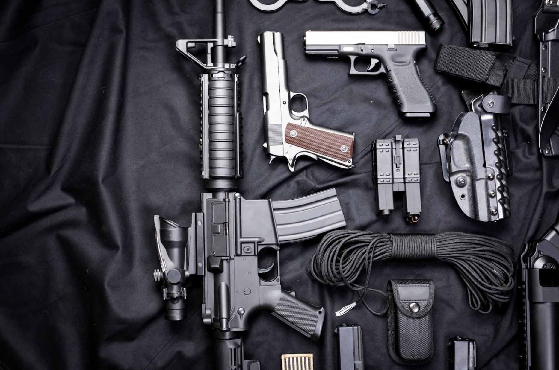 A collection of firearms and accessories displayed on a black cloth: an Armalite rifle, two types of revolvers, a handcuff, and various accessories.