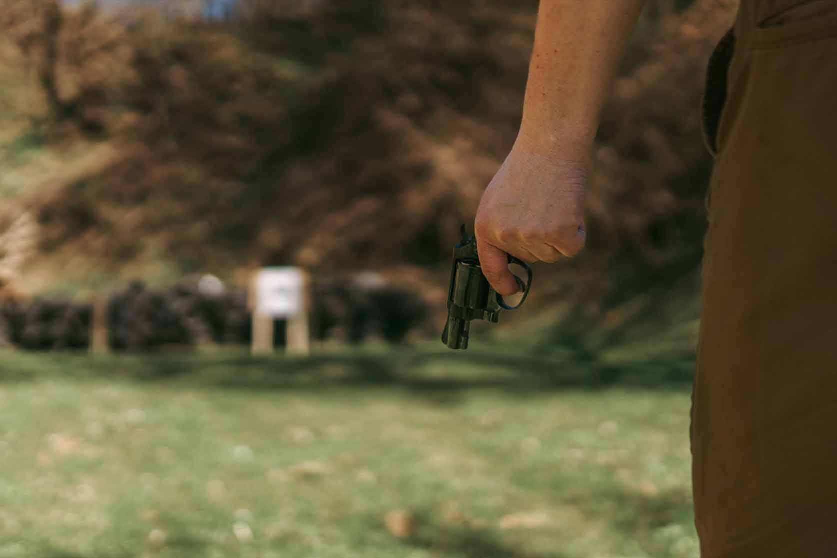 A male gun enthusiast holds a revolver, ready to aim at a distant target on an outdoor shooting range; identity obscured.