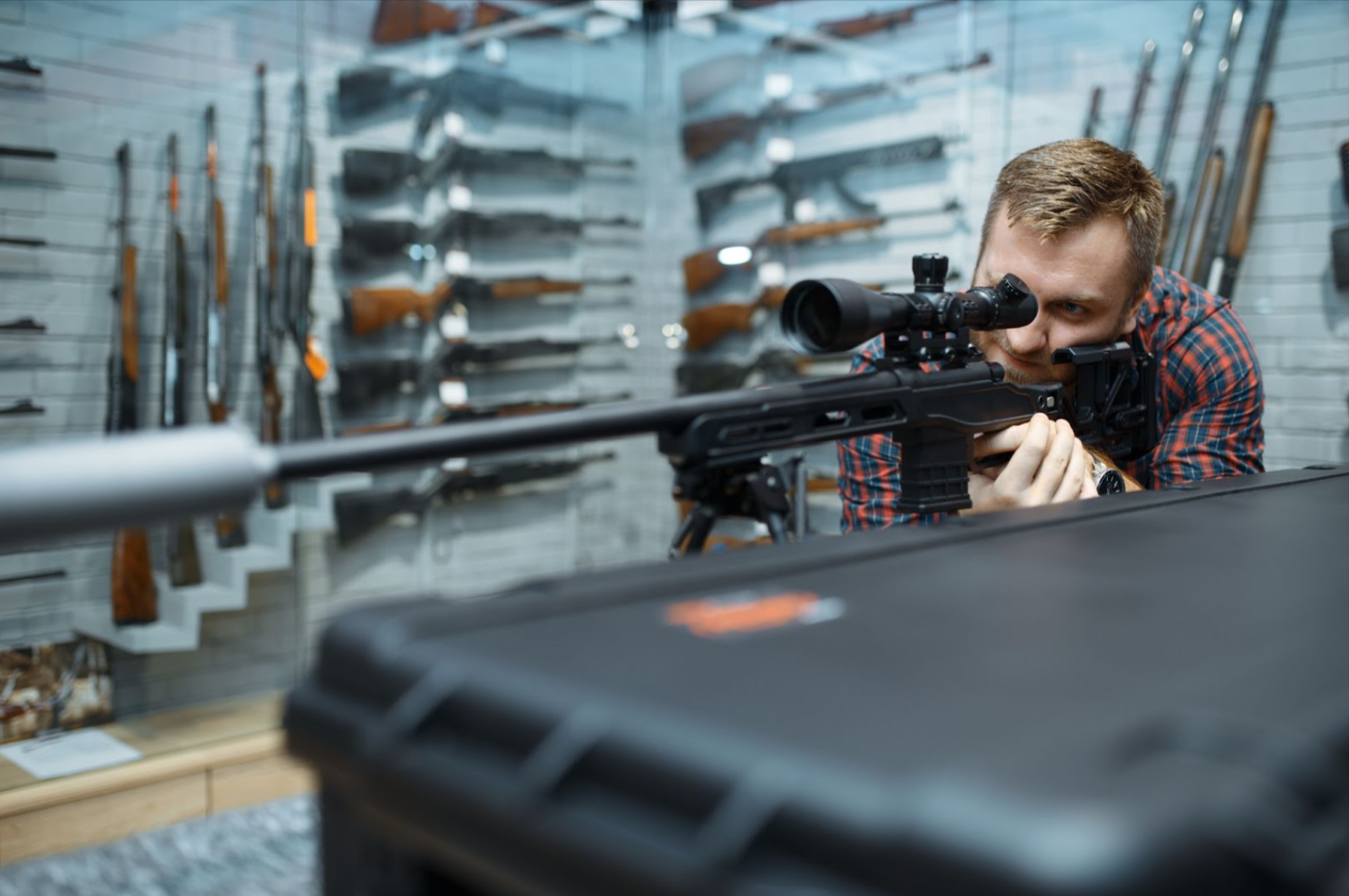 A man aims with a sniper rifle in a gun shop, surrounded by an array of rifles on display in the background.