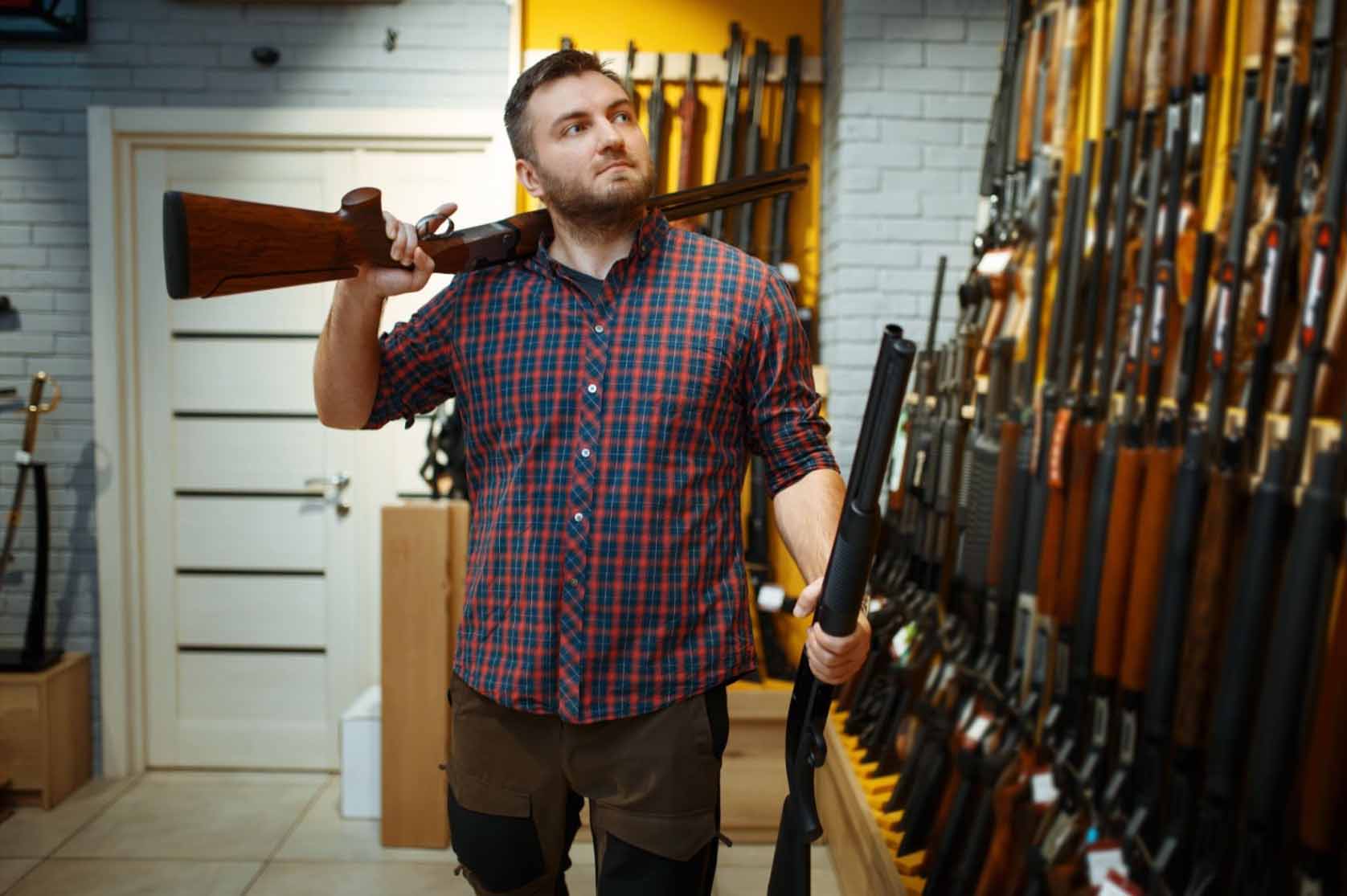 A man holds rifles in both hands while looking at the display and contemplating his choice at a gun shop.