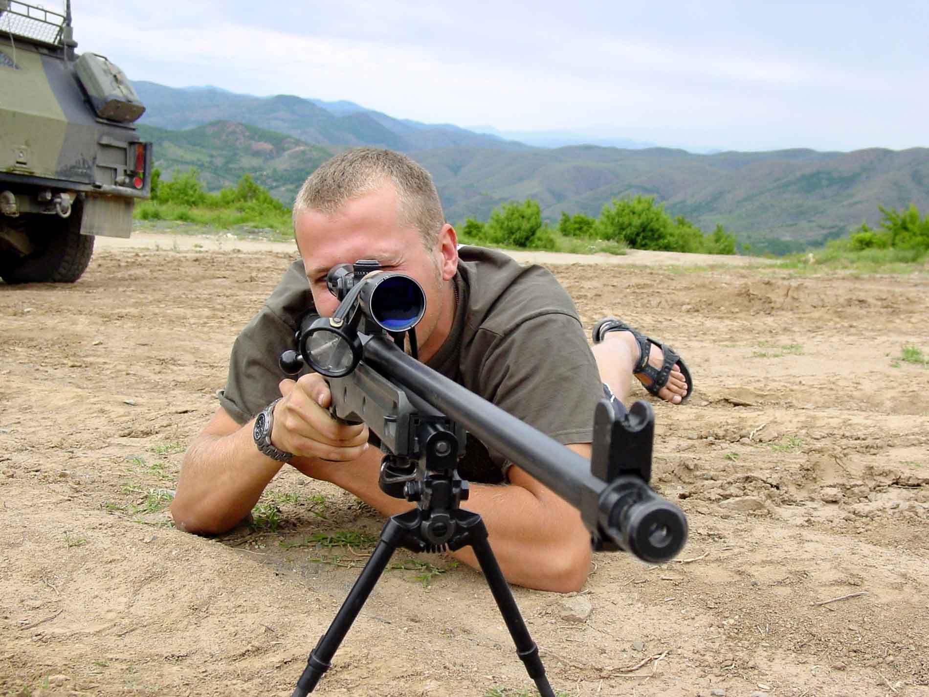 A man looking through the sight of a black sniper rifle on a gun bipod.