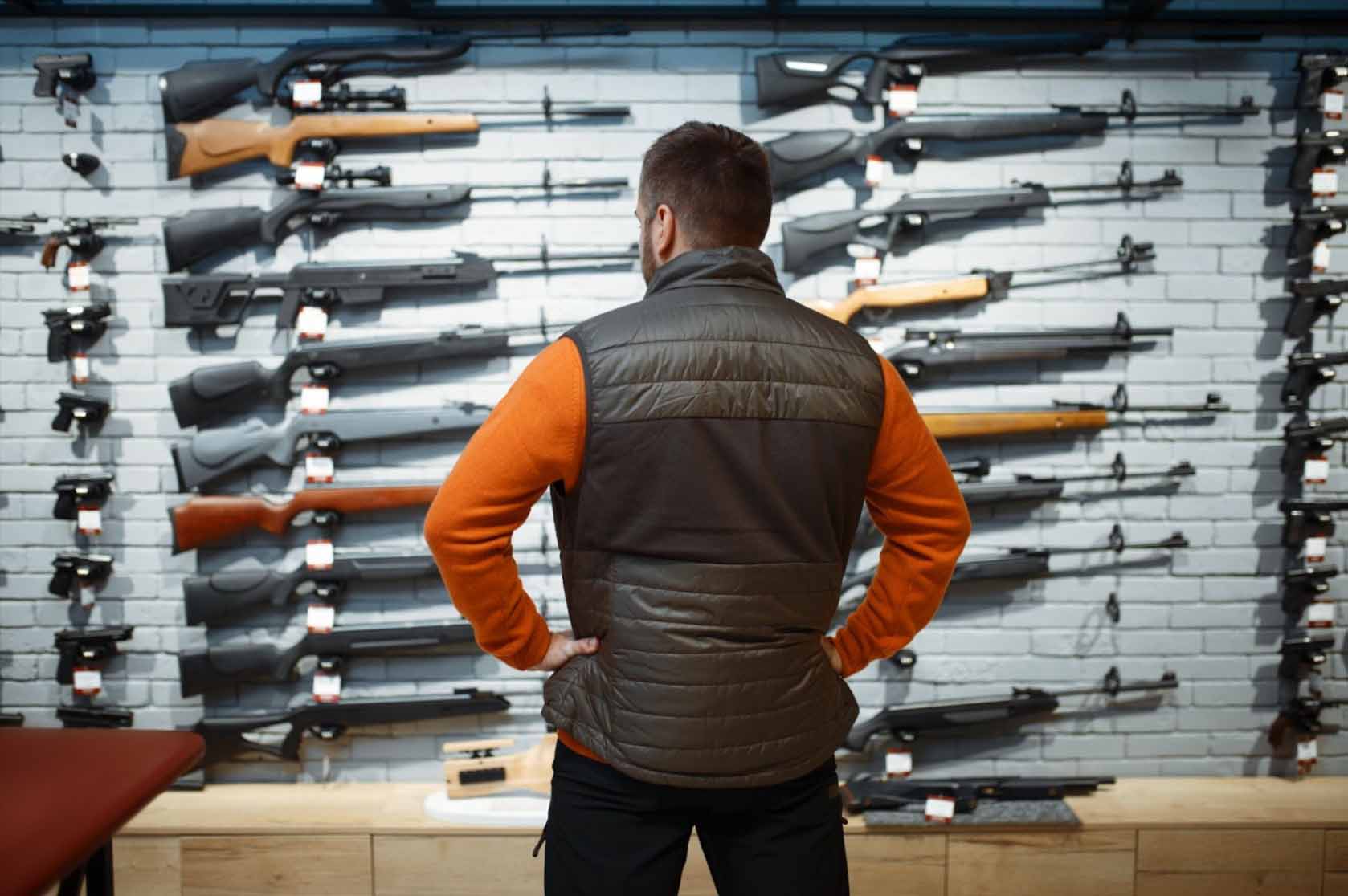 A man views a rifle display case in a gun shop, contemplating the selection.