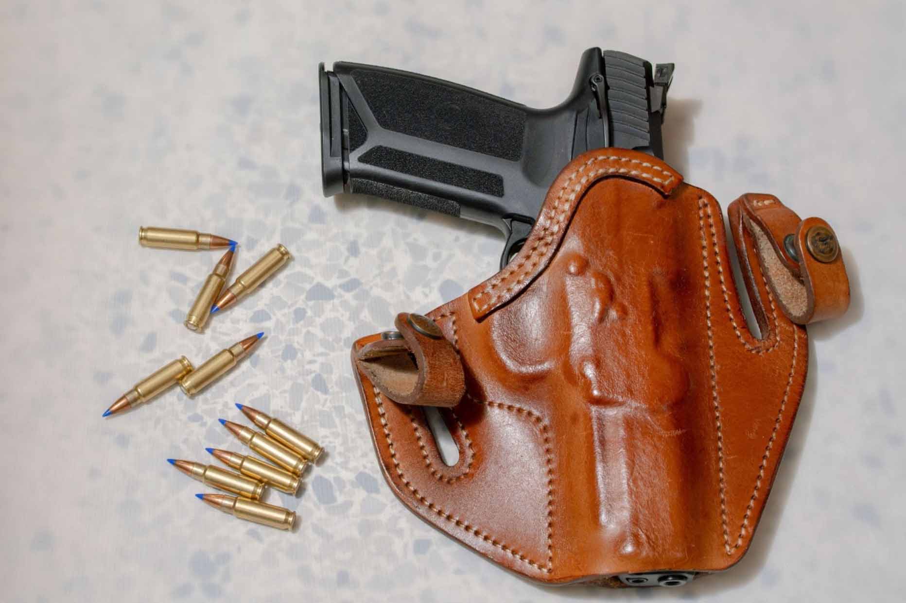 A revolver in a brown holster is placed on a white background, with loose bullets on the left side.