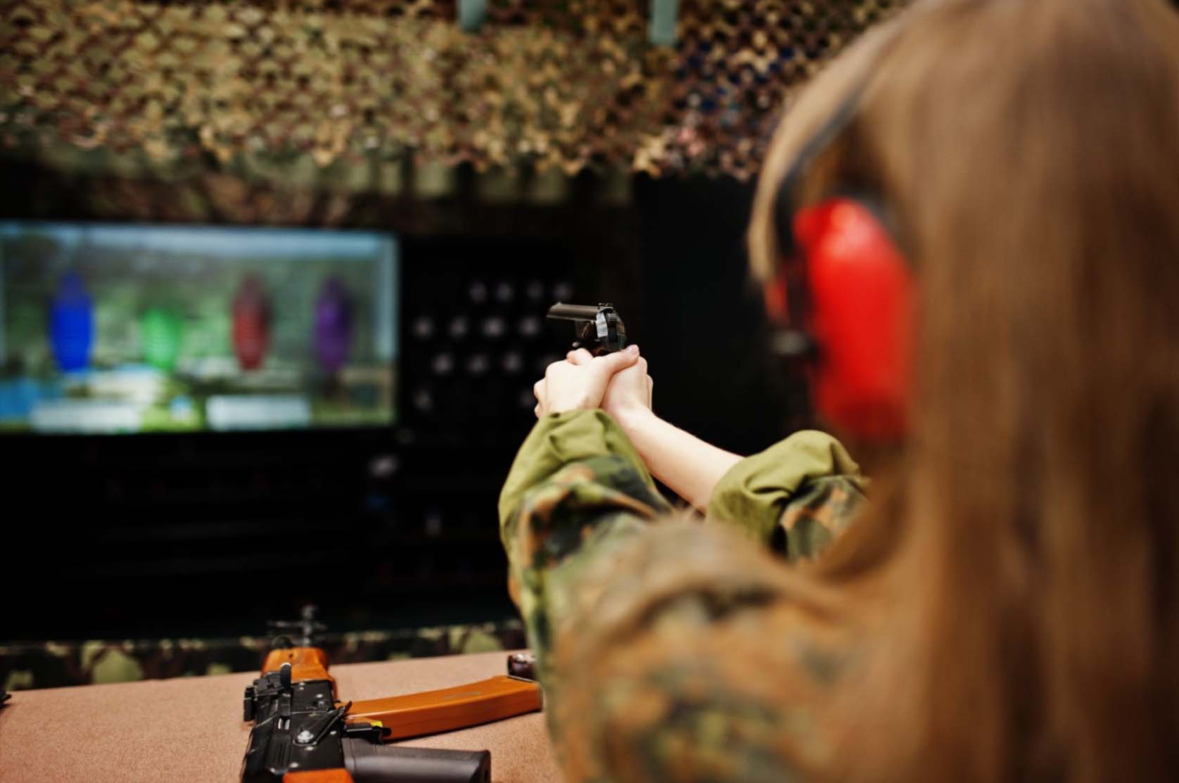 A woman wearing a vibrant red protective headset is seen from behind as she aims her firearm at water jugs at a shooting range.