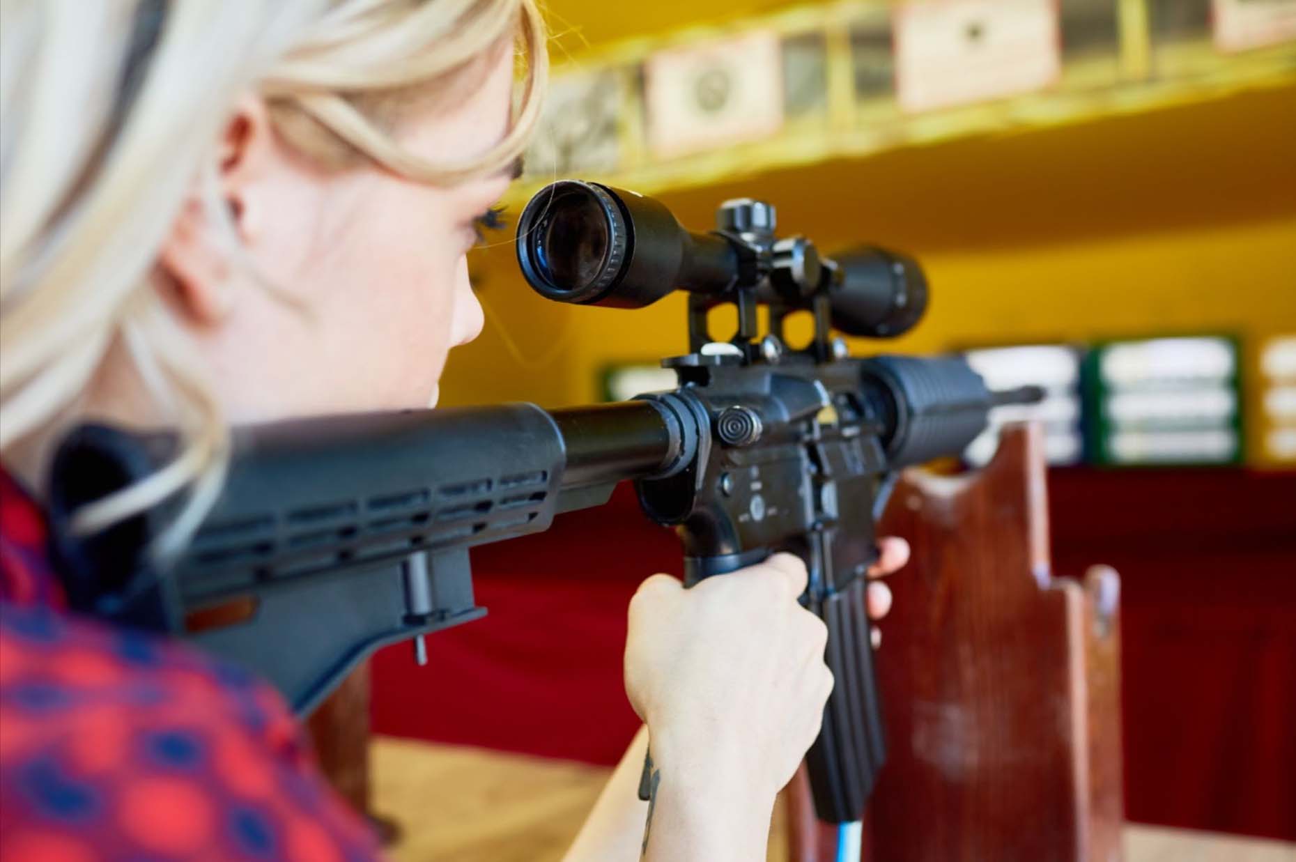 A woman wearing flannel stares down the barrel of a rifle at a shooting range.