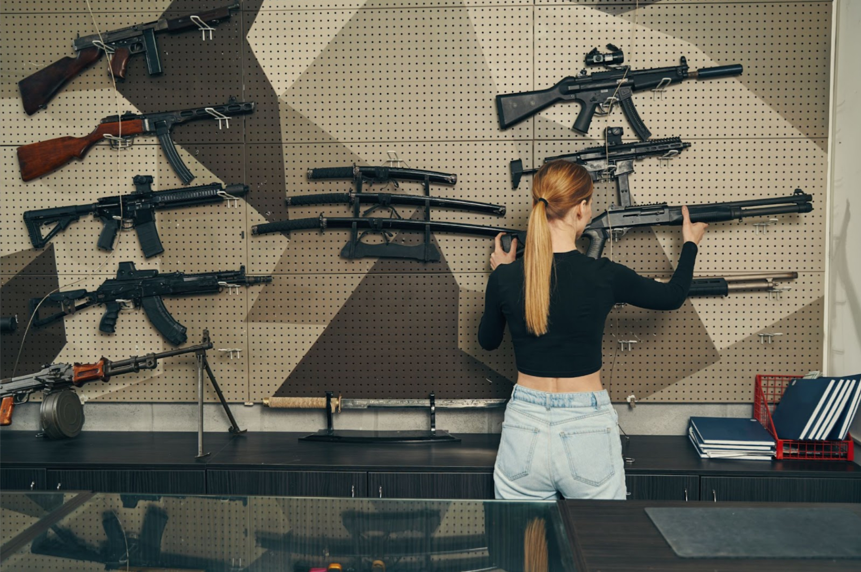 An experienced blonde saleswoman, with her back to the camera, selects a rifle from the display.