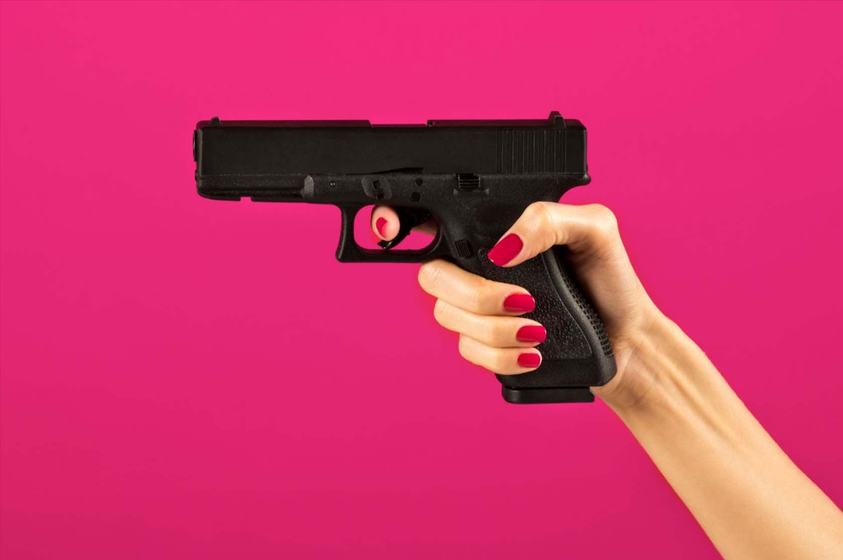Close-up of a woman's hand with red nail polish gripping a pistol, set against a vibrant pink background.