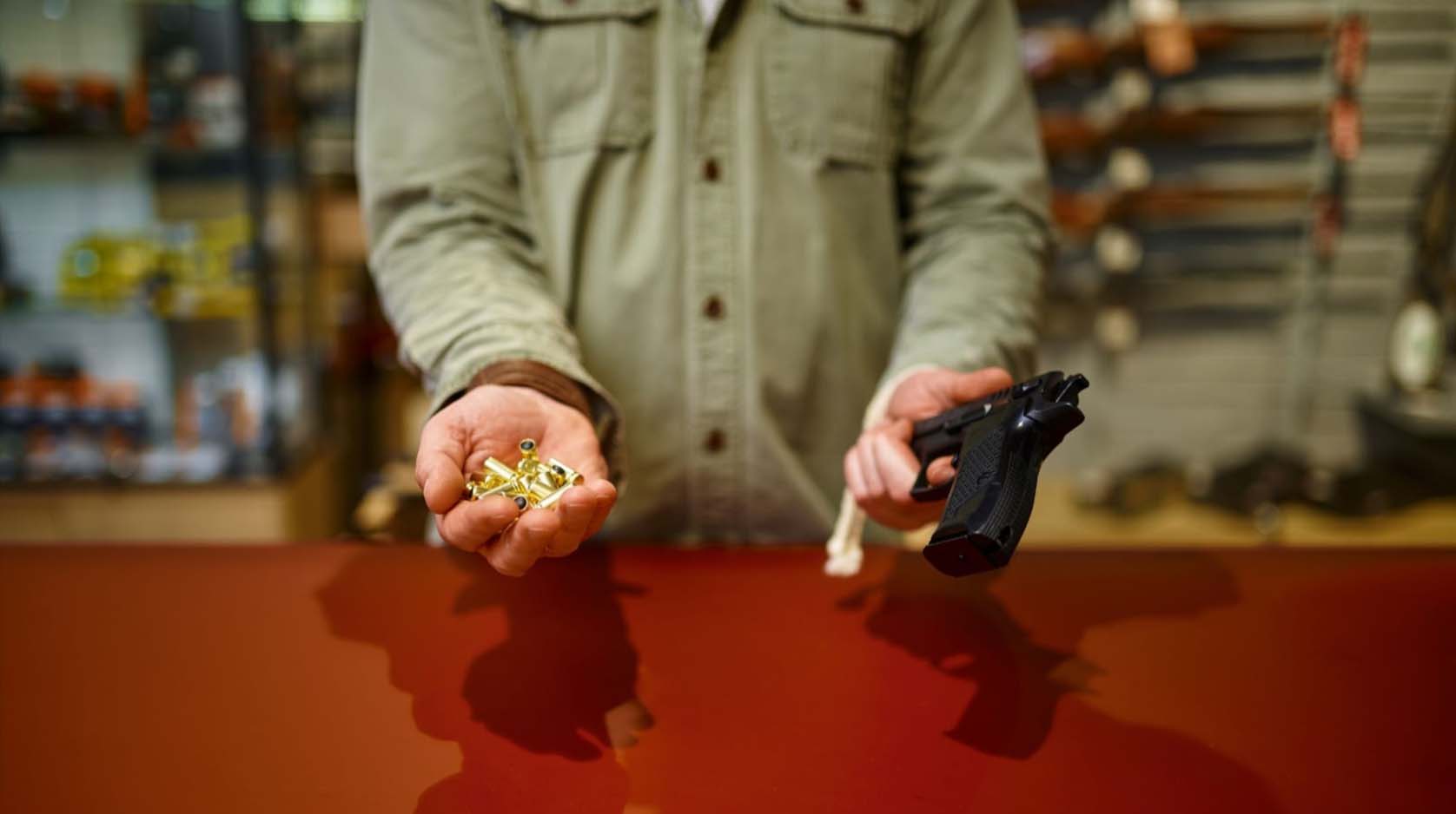 Close-up view of a gun store clerk’s hand presenting a handgun and ammunition to a customer.