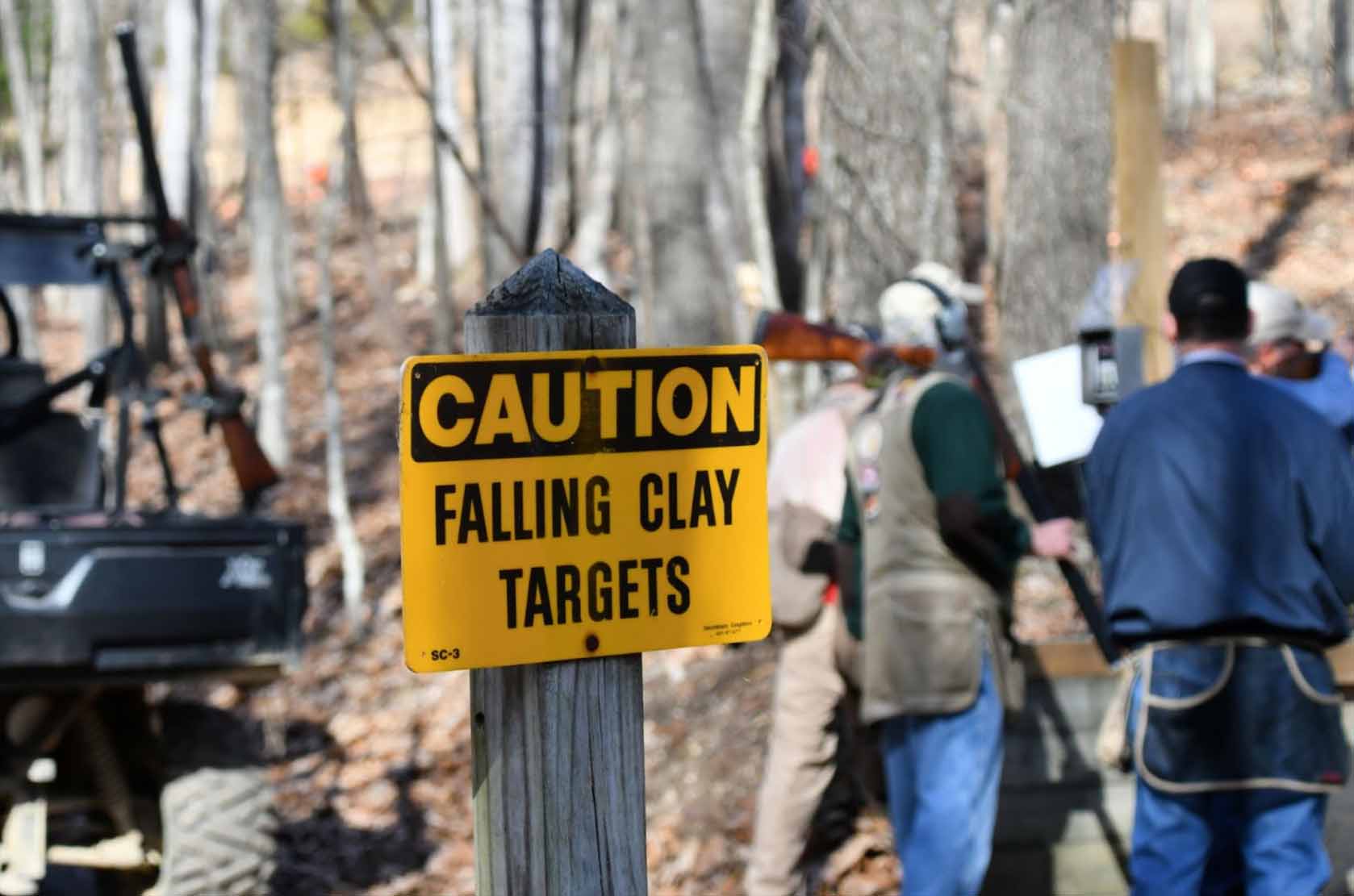 Caution: Falling Clay Targets sign with a blurred background of men holding shotguns at a shooting range.