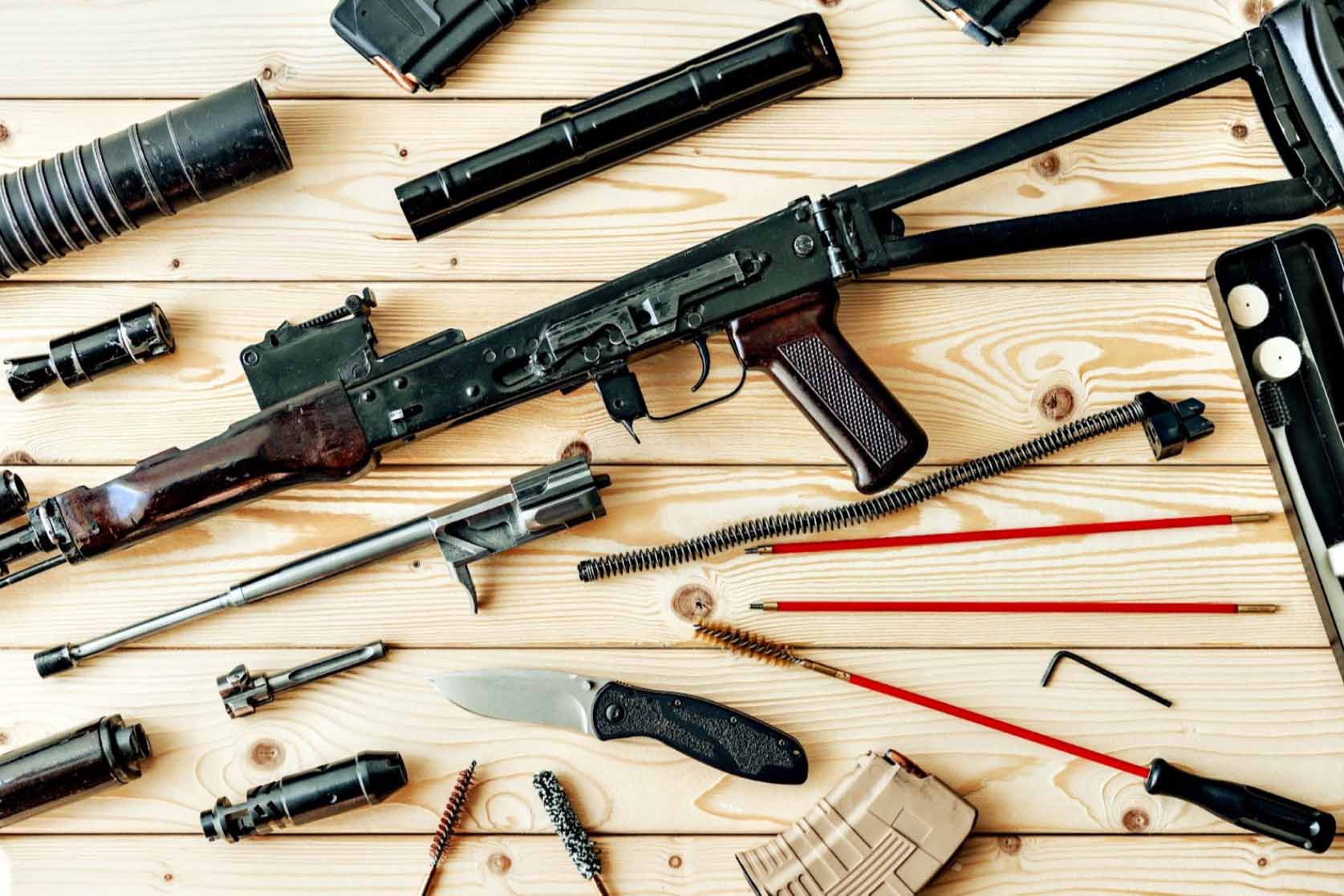 In the close-up shot, a disassembled rifle rests on a table in a weapons workshop, surrounded by various cleaning tools.