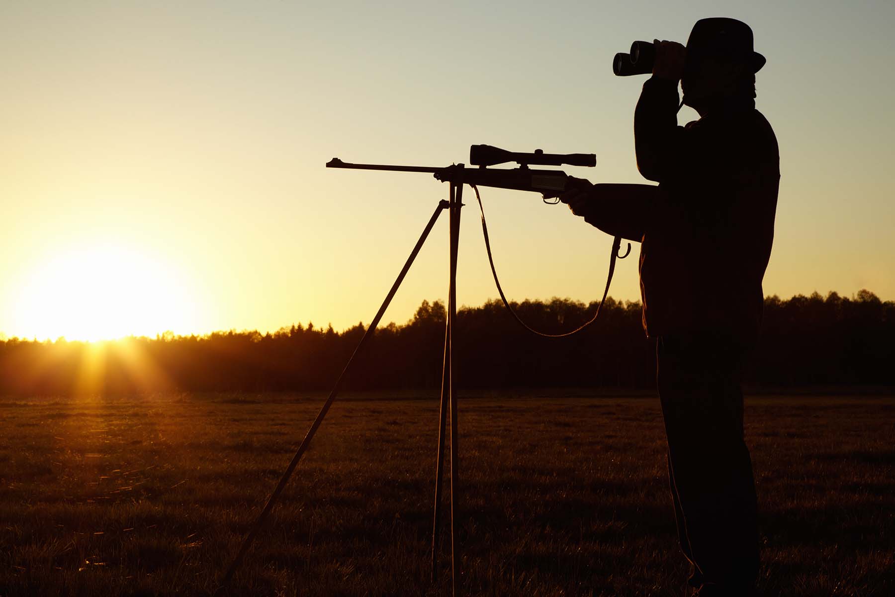 The silhouette of a man wearing a hat and looking through binoculars holds a rifle on top of a tripod.