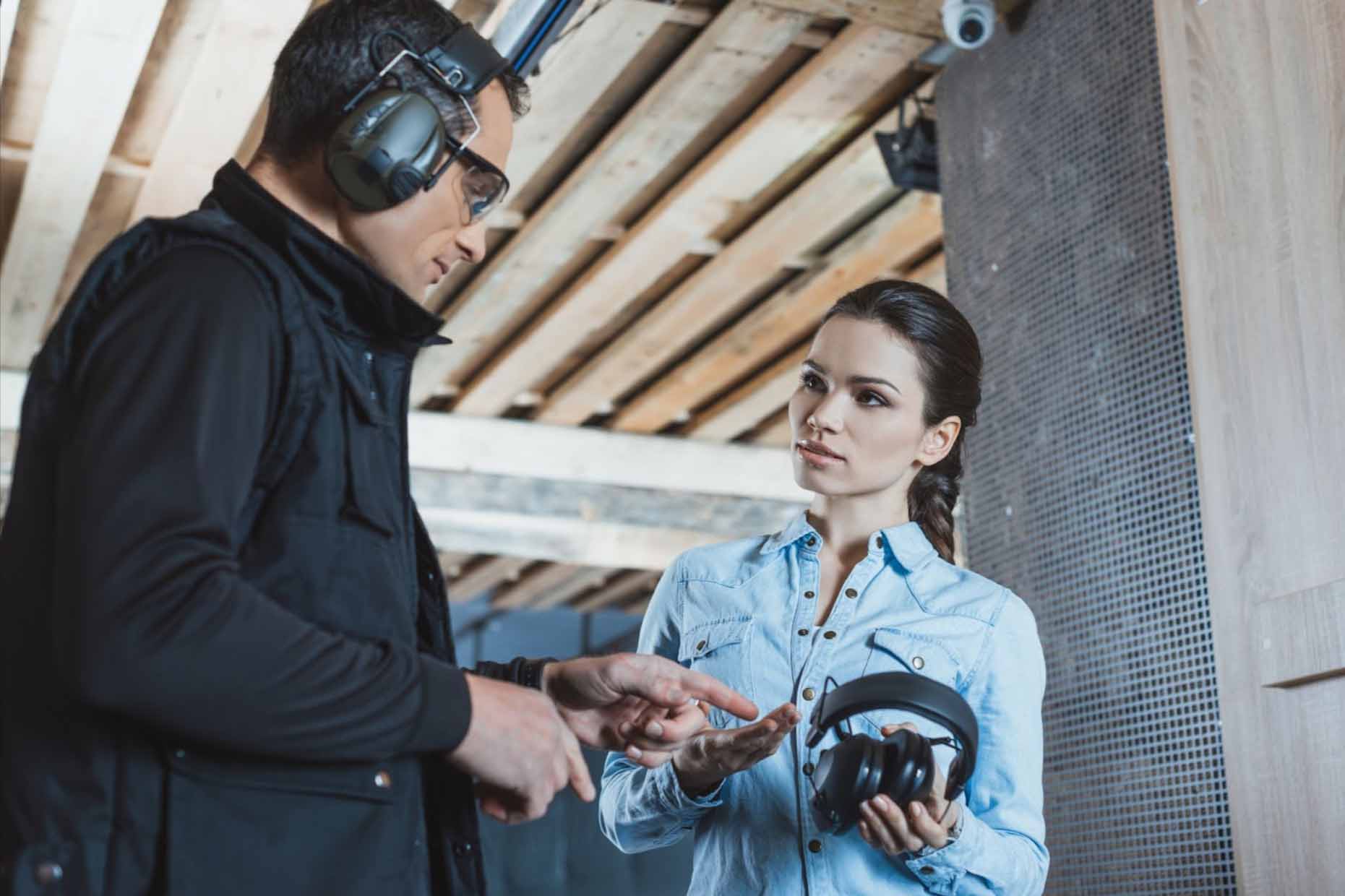 Woman holding headphones next to a male instructor while at a gun range.